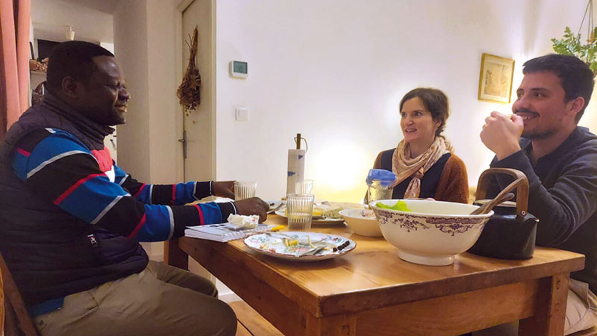 Un homme est assis à table face à un couple, qui l’écoutent, devant des assiettes vides et un saladier.