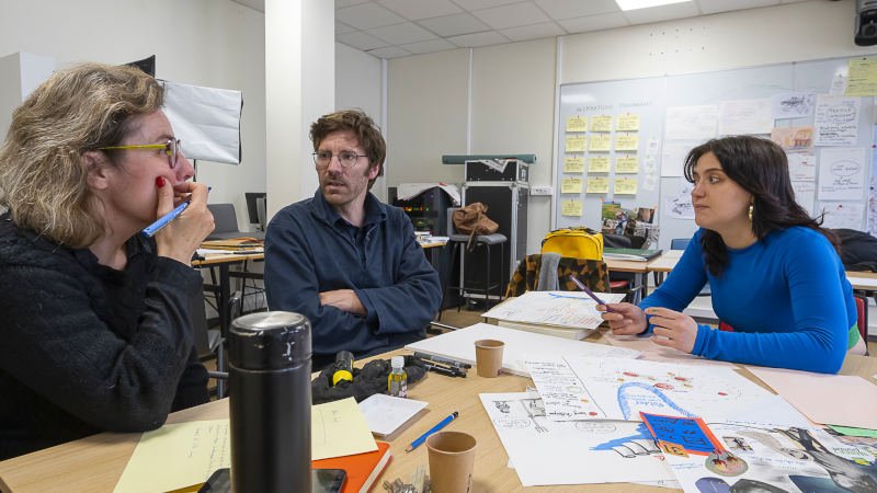 2 femmes et en homme en discussion, environnés de feuilles, croquis, postits, gobelets, gourde…