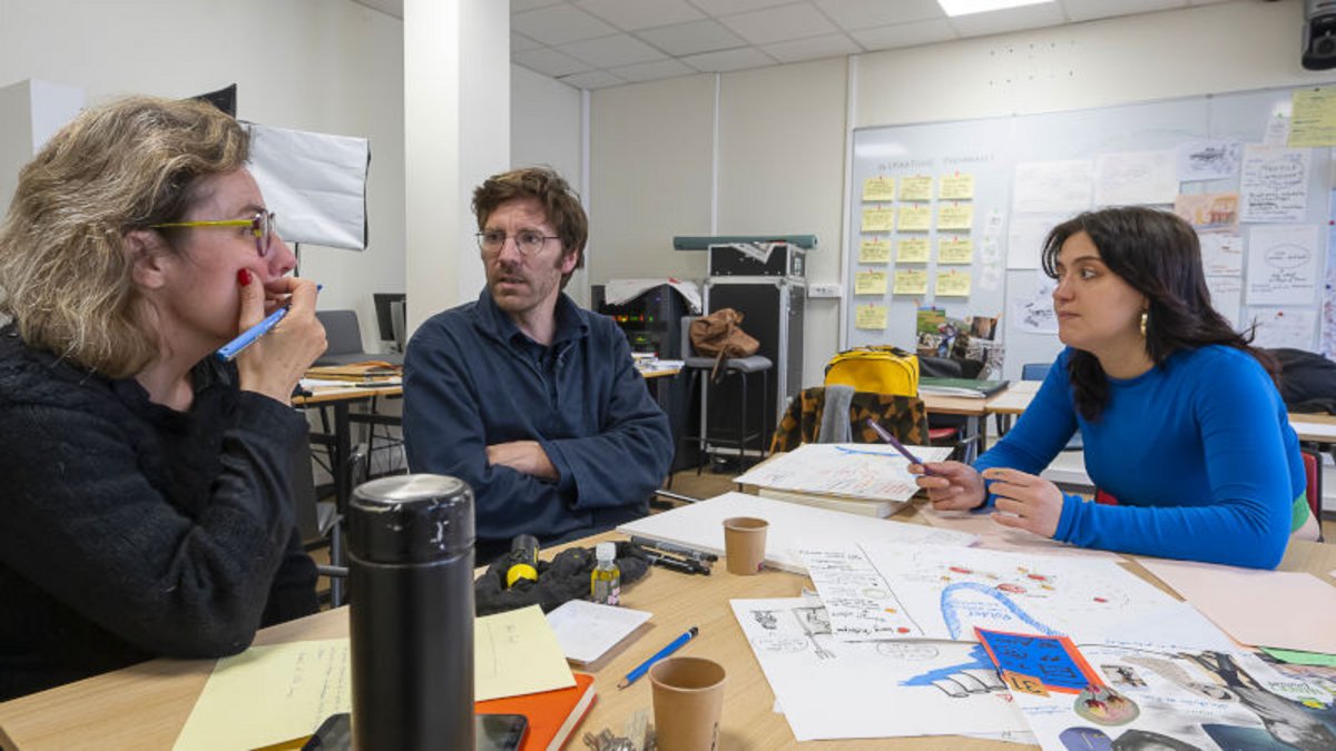 2 femmes et en homme en discussion, environnés de feuilles, croquis, postits, gobelets, gourde…
