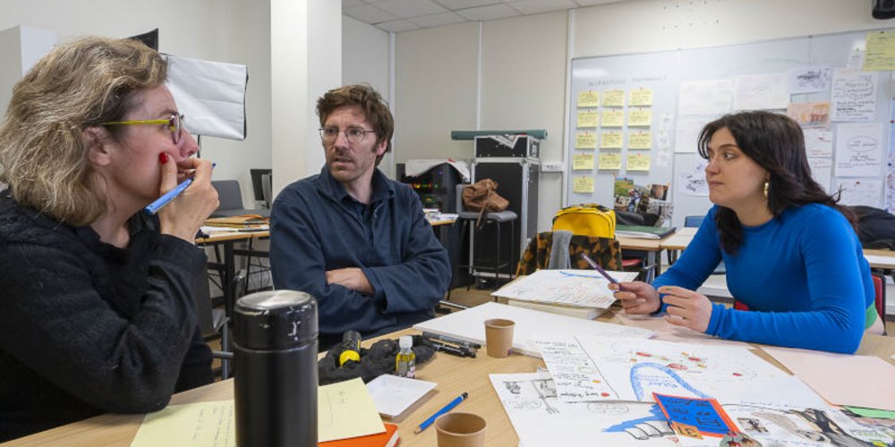 2 femmes et en homme en discussion, environnés de feuilles, croquis, postits, gobelets, gourde…