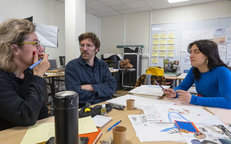 2 femmes et en homme en discussion, environnés de feuilles, croquis, postits, gobelets, gourde…