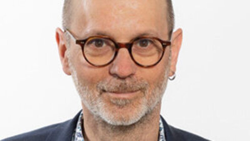 Un homme avec une veste, des lunettes, une boucle d’oreille et une chemise fleurie, regarde l’objectif en souriant.