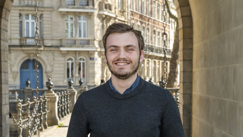 Un jeune homme souriant se tient devant une ouverture type porte cochère, en arrière-plan des immeubles en pierre