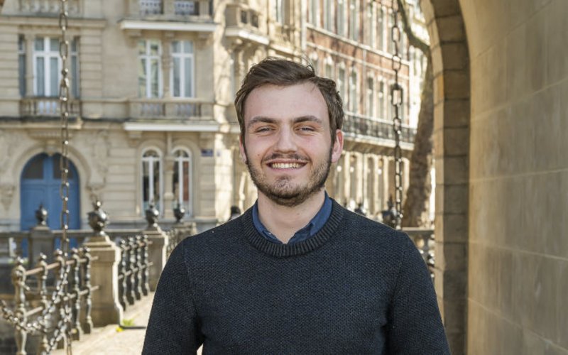 Un jeune homme souriant se tient devant une ouverture type porte cochère, en arrière-plan des immeubles en pierre