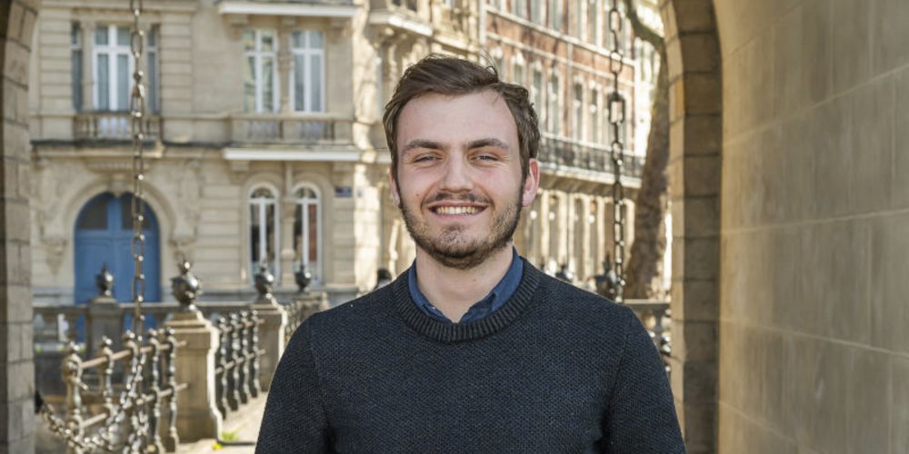 Un jeune homme souriant se tient devant une ouverture type porte cochère, en arrière-plan des immeubles en pierre
