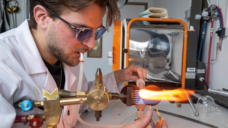 Un jeune homme avec des lunettes teintées, souffle, tient un tube dans sa main gauche qu’il passe au chalumeau, son autre main le manipulant avec des pincettes. 