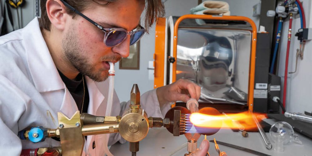 Un jeune homme avec des lunettes teintées, souffle, tient un tube dans sa main gauche qu’il passe au chalumeau, son autre main le manipulant avec des pincettes. 
