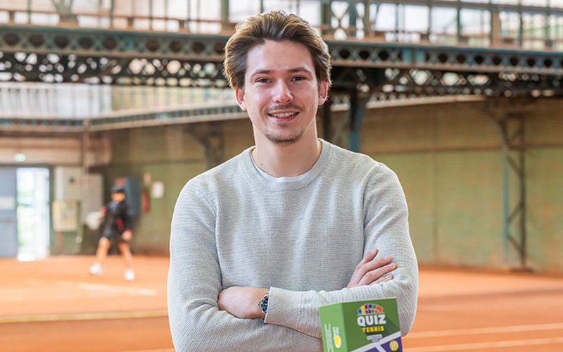 Un jeune homme souriant et bras croisés, avec une boîte sur laquelle est écrit « Quiz ». Dans le fond, un court couvert en terre battue.