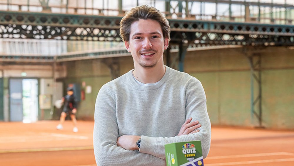 Un jeune homme souriant et bras croisés, avec une boîte sur laquelle est écrit « Quiz ». Dans le fond, un court couvert en terre battue.