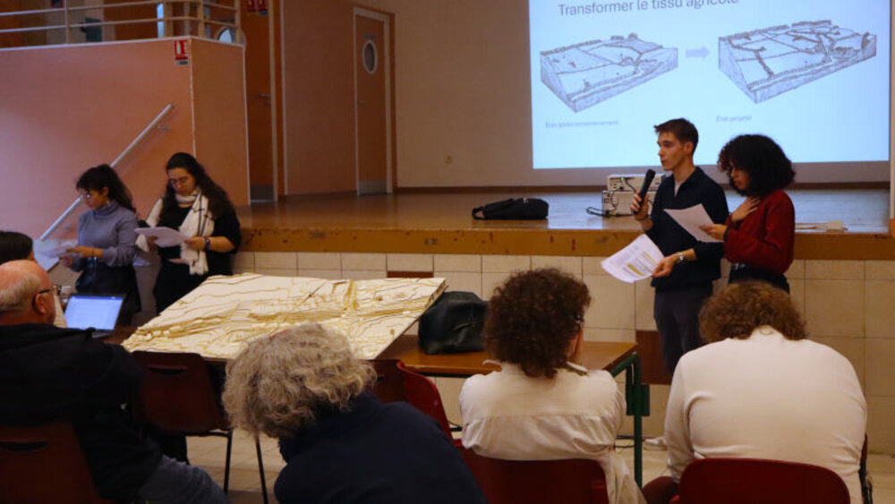 Un jeune homme et une jeune fille au micro, un dessin de terrain agricole derrière eux, et une maquette devant. Assis, des habitants.