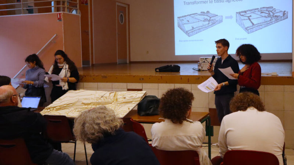 Un jeune homme et une jeune fille au micro, un dessin de terrain agricole derrière eux, et une maquette devant. Assis, des habitants.