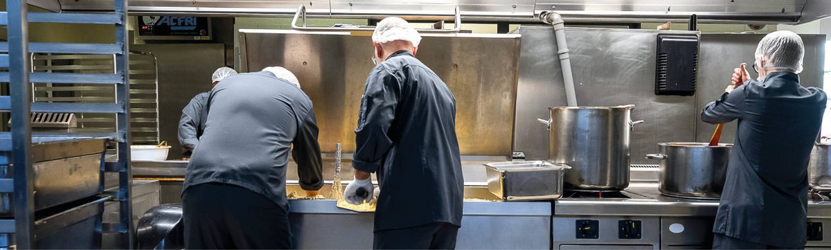 Deux cuisiniers à gauche et une cuisinière à droite, s’affairent dans un environnement de cuisine inox. On distingue deux marmites.