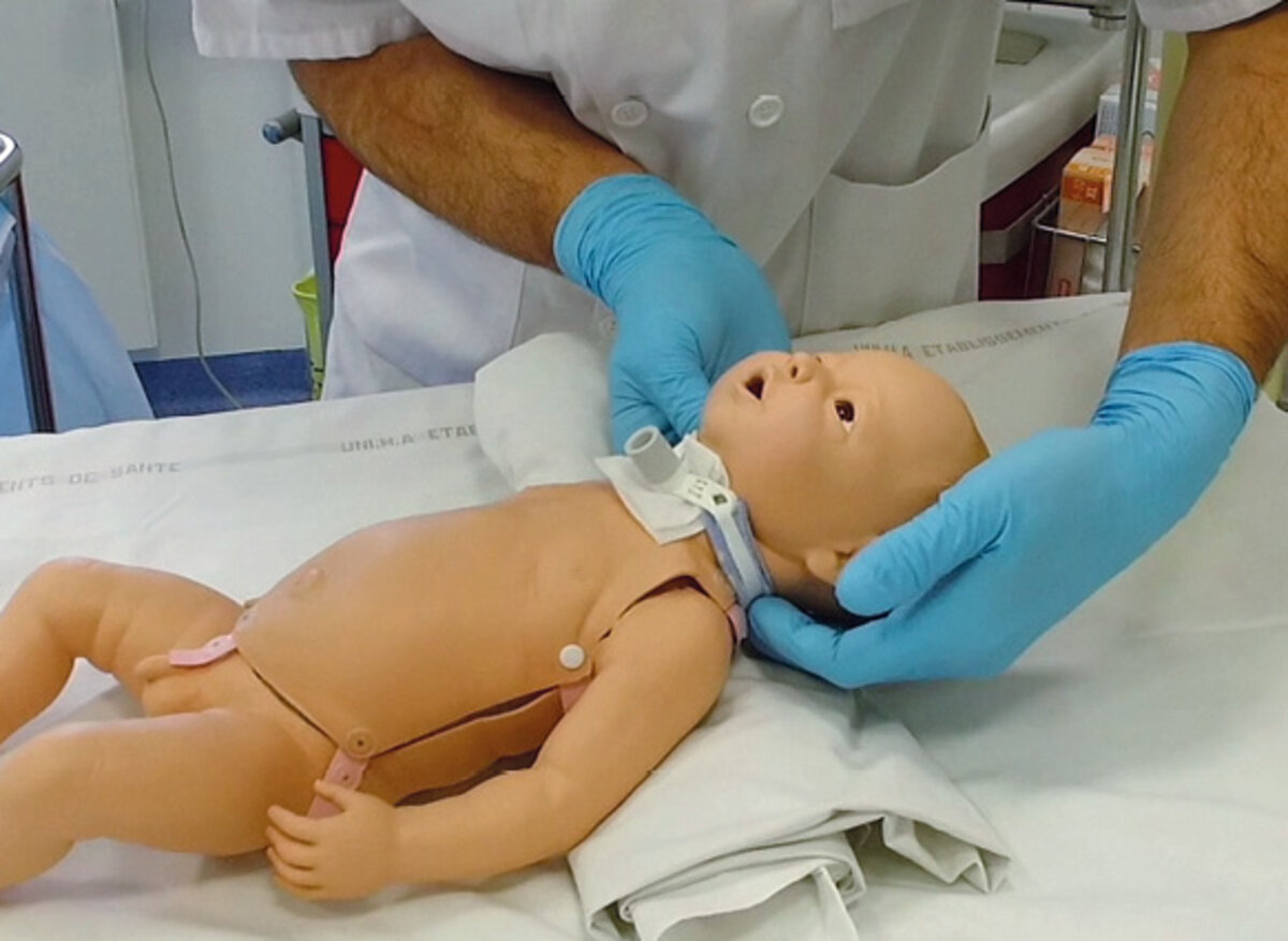 Mannequin de bébé, avec sur le cou un dispositif, manipulé par un homme avec des gants bleus.
