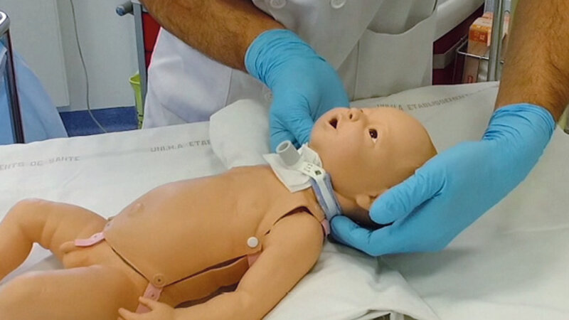 Mannequin de bébé, avec sur le cou un dispositif, manipulé par un homme avec des gants bleus.