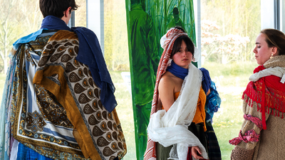 Un jeune homme de dos, avec une cape colorée et avec des motifs, fait face à deux jeunes filles avec des costumes similaires. En arrière-plan, une peinture de dominante verte.