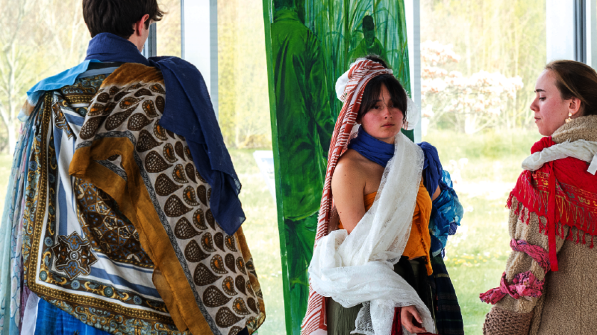 Un jeune homme de dos, avec une cape colorée et avec des motifs, fait face à deux jeunes filles avec des costumes similaires. En arrière-plan, une peinture de dominante verte.