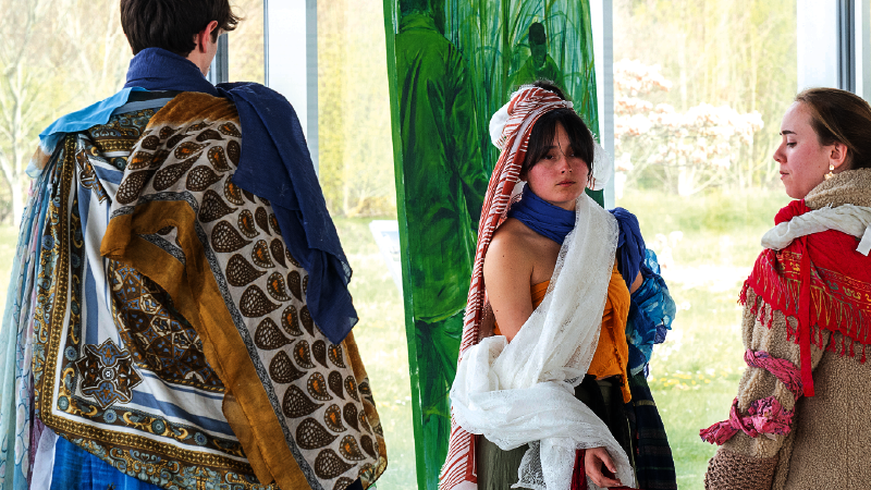 Un jeune homme de dos, avec une cape colorée et avec des motifs, fait face à deux jeunes filles avec des costumes similaires. En arrière-plan, une peinture de dominante verte.