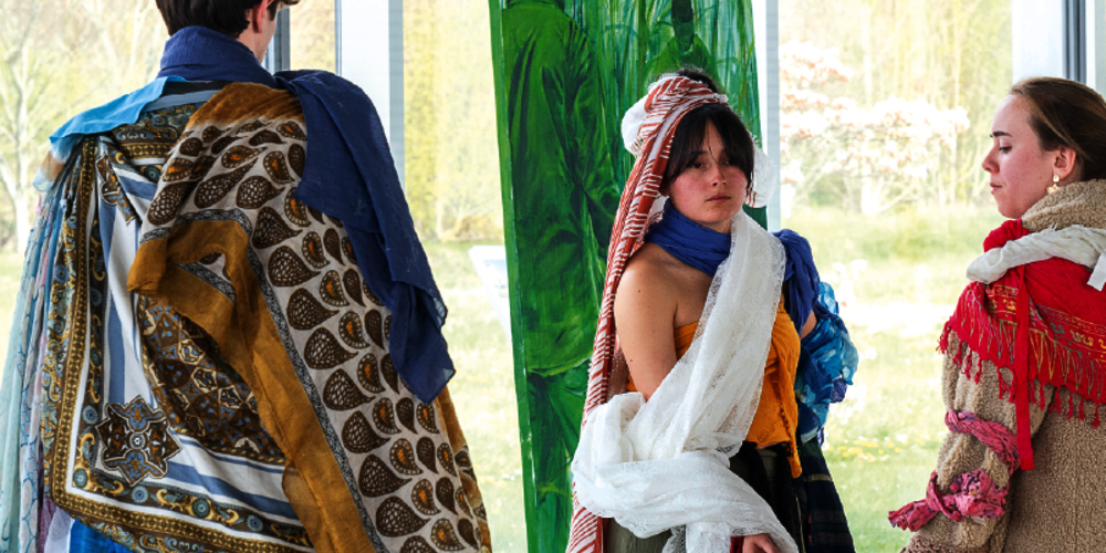 Un jeune homme de dos, avec une cape colorée et avec des motifs, fait face à deux jeunes filles avec des costumes similaires. En arrière-plan, une peinture de dominante verte.