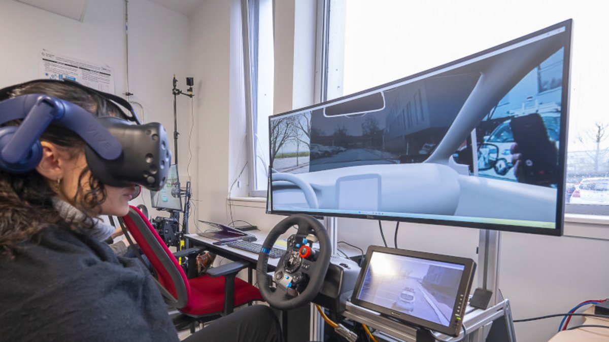 Une jeune femme avec un casque VR, face à un écran séparé en 2 (vue réelle/virtuelle), avec un volant en-dessous.