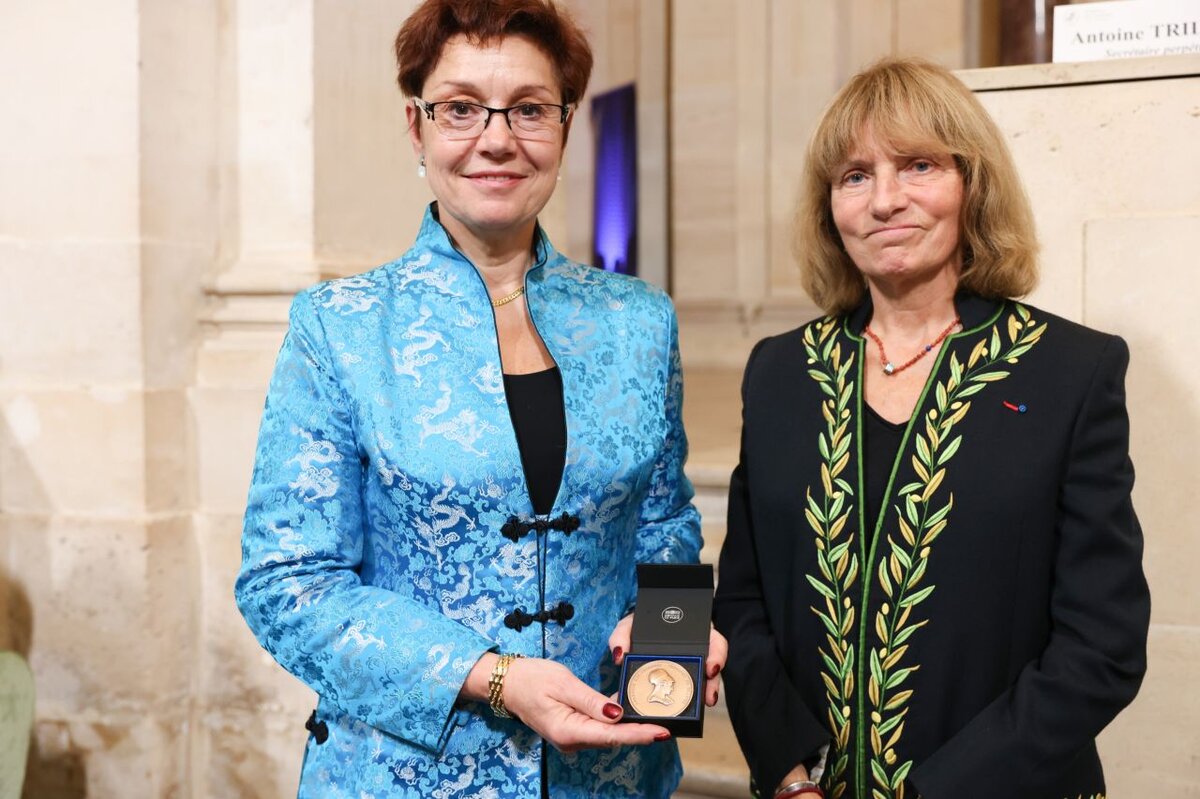 Une femme habillé en bleue, montre une médaille à côté d’une autre femme en habit d’académicienne