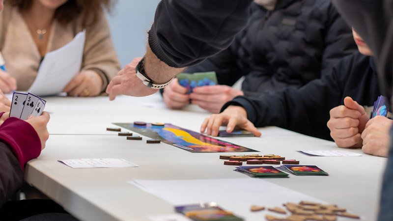 Un jeu de plateau sur une table, avec des cartes et des jetons. Dans les mains d’un des participants, des cartes à jouer.