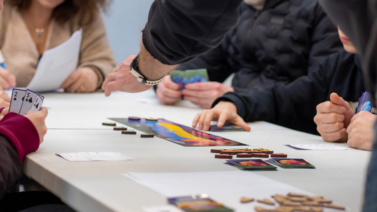 Un jeu de plateau sur une table, avec des cartes et des jetons. Dans les mains d’un des participants, des cartes à jouer.