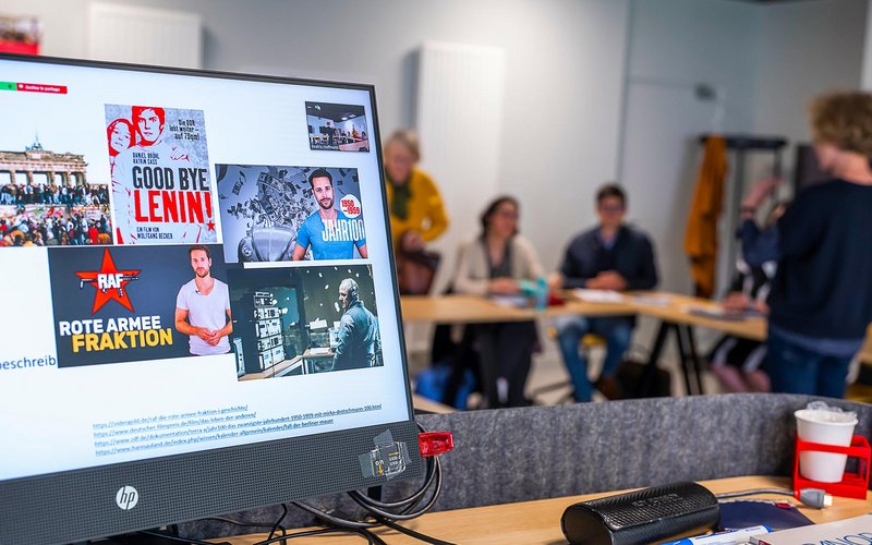 Un écran avec plusieurs images dont l’affiche du film Goodbye Lenin, et dans le fond les participants au cours, assis, et l’enseignante, debout