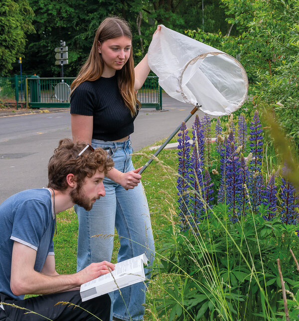 Devant des plantes, un jeune homme, accroupi, consulte un manuel, tandis qu’une jeune fille, debout, tient une sorte de filet à papillon