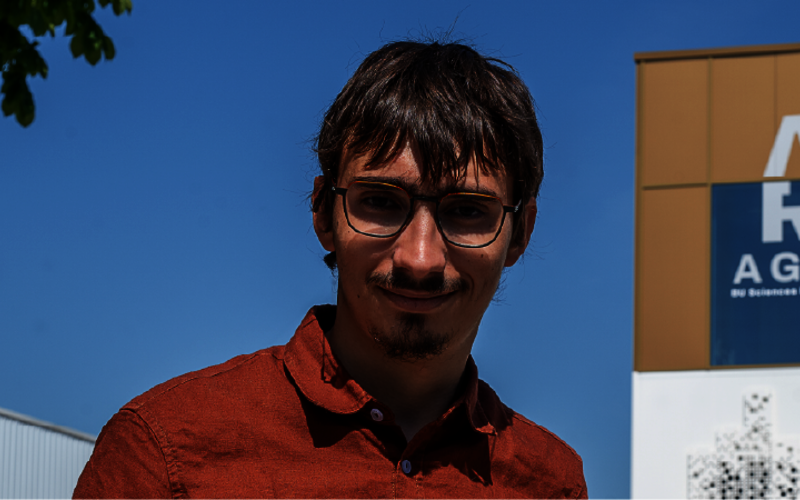 Jeune homme avec bouc et lunettes, en chemise ocre rouge. À sa gauche, un bâtiment neuf.