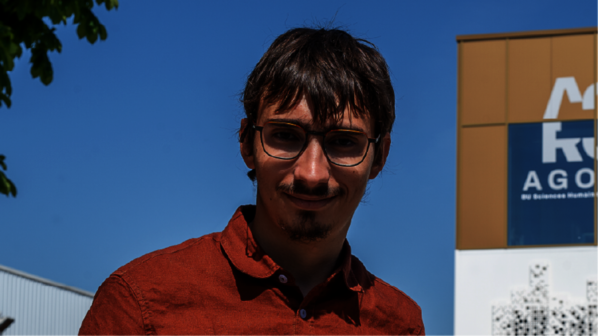 Jeune homme avec bouc et lunettes, en chemise ocre rouge. À sa gauche, un bâtiment neuf.