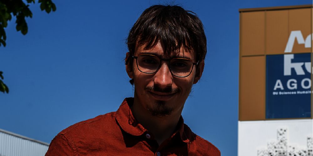 Jeune homme avec bouc et lunettes, en chemise ocre rouge. À sa gauche, un bâtiment neuf.