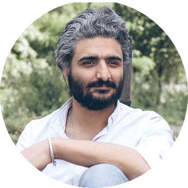 Un homme en chemise blanche, barbu, les cheveux poivre et sel, avec de la végétation derrière lui.