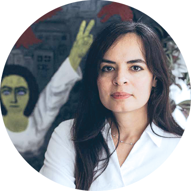 Une femme avec une chemise blanche, regarde l’objectif. Derrière elle, une peinture avec une jeune fille les bras levés.