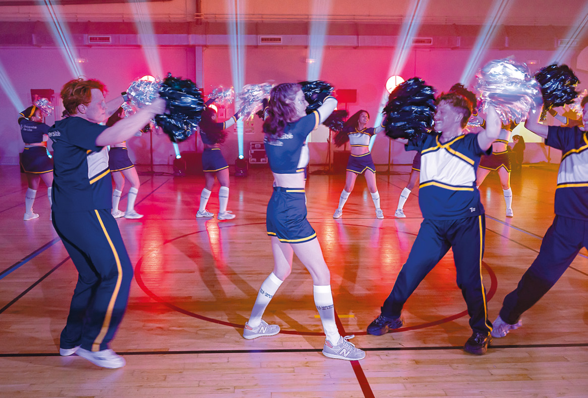 Des jeunes hommes et jeunes femmes agitent des grands pompons, sous une lumière rose-orangée