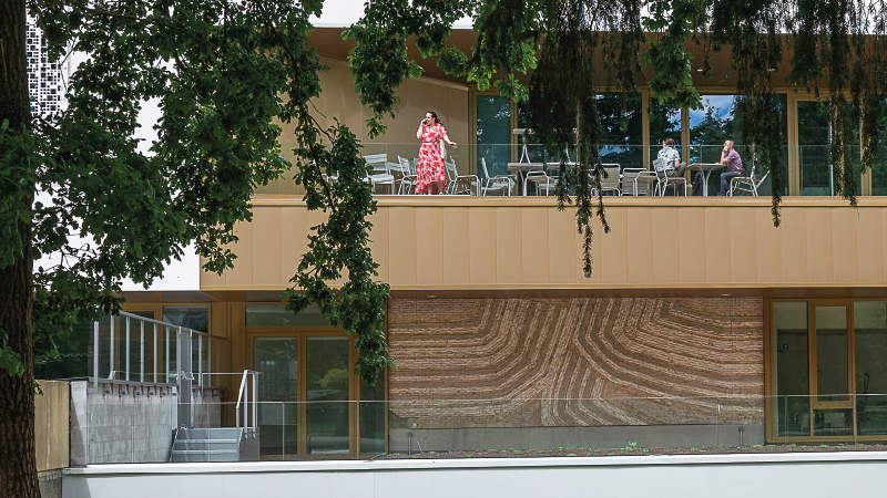 Terrasse sur deux étages sur la façade d’un bâtiment. En haut, des personnes attablées. En bas, une fresque murale avec des traits parallèles. Au premier plan, les feuilles et le tronc d’un arbre.