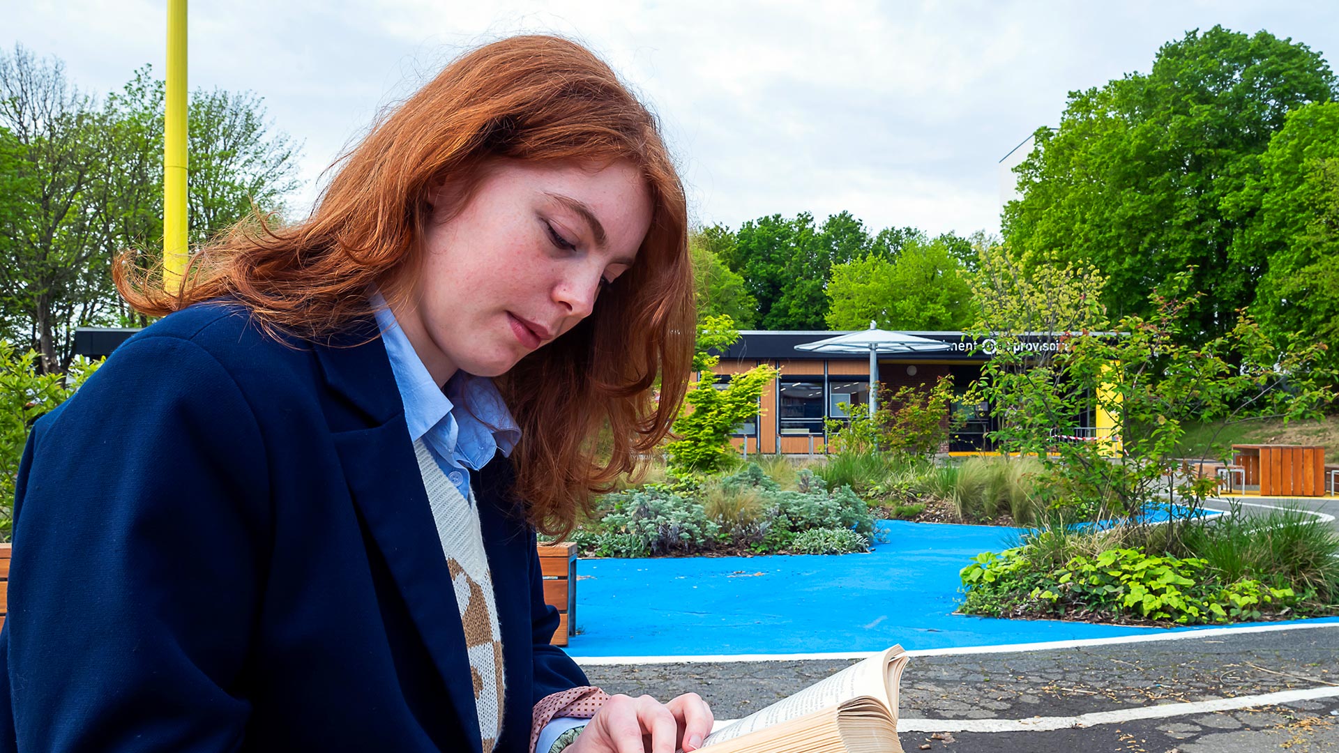 Une jeune fille lit un livre, en arrière-plan un sol bleu, des arbustes et un bâtiment préfabriqué au loin